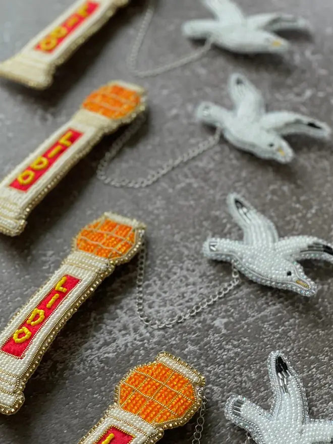 Beaded lido brooch with orange, red, and beige beads attached to a white beaded seagull by a silver chain, displayed on a grey table. 