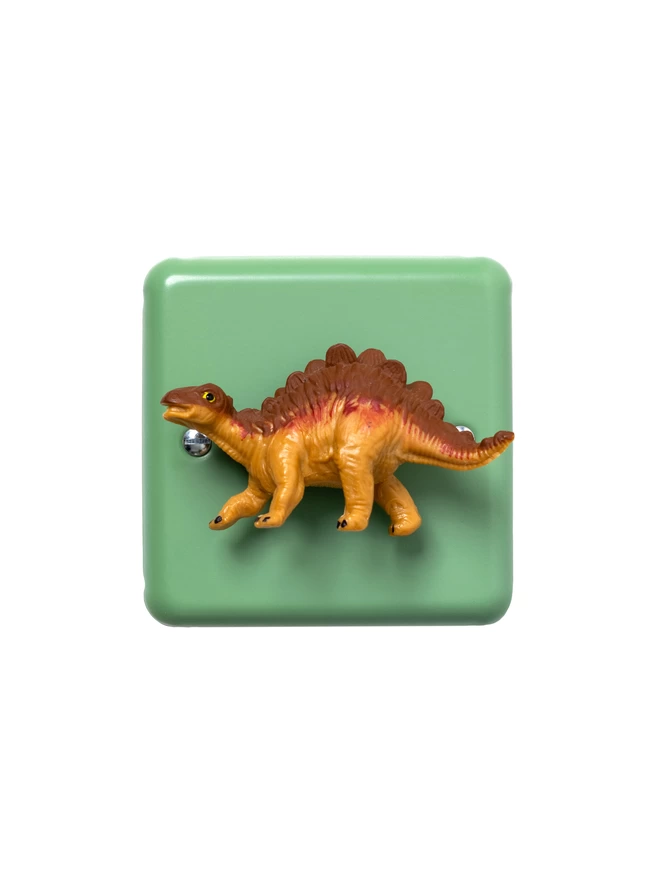A Stegosaurus dinosaur light switch. The light switch has a pastel green steel metal epoxy coated light switch plate, the Stegosaurus dinosaur is orange and made of plastic. The light switch is made by Candy Queen Designs and Varilight.