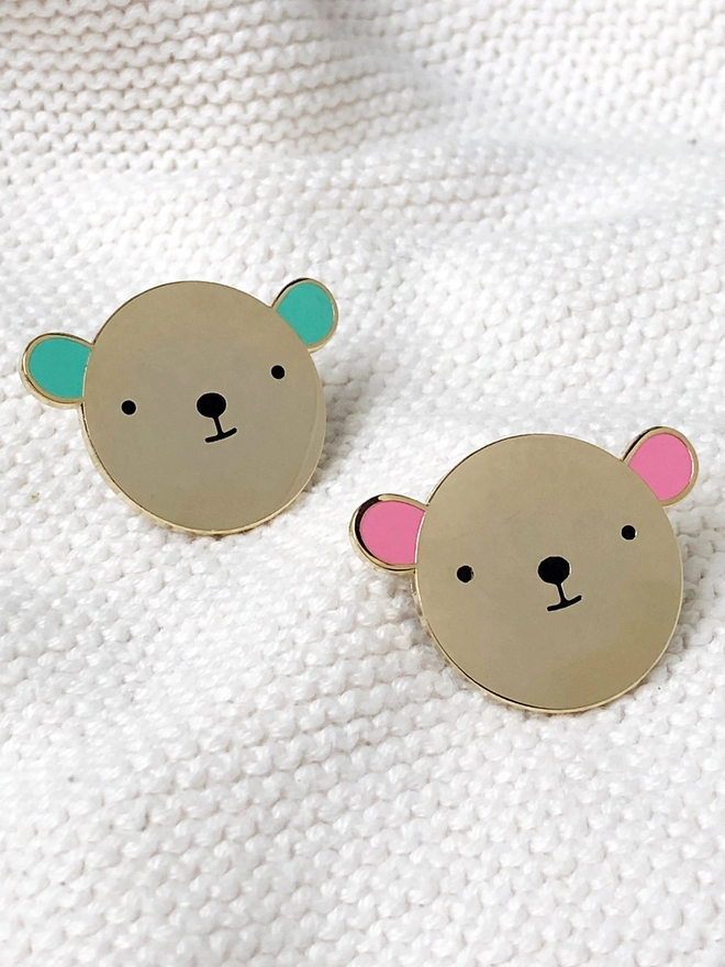 Two gold bear enamel pins, one with green ears and one with pink ears, are resting on white fabric.
