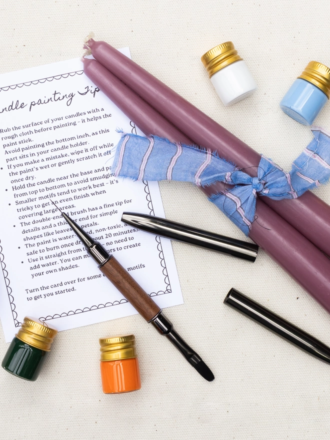 Flat lay of a candle painting kit with two mauve taper candles, a double-ended paintbrush, six colourful pots of candle paint and an illustrated tips card on a neutral fabric background.
