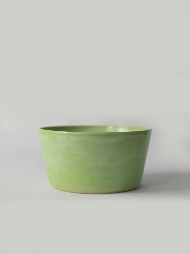 A single hand-thrown ceramic breakfast bowl in spring green, with a glossy glaze showing subtle variation in tone, a gently flared form and unglazed natural clay base, photographed against a plain light grey background.