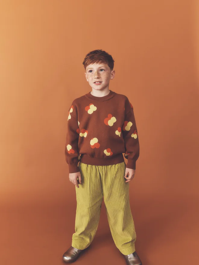 Close crop of a child with hands in the pockets of chartreuse wide-leg corduroy trousers, worn with a Studio Koter chestnut floral intarsia jumper.