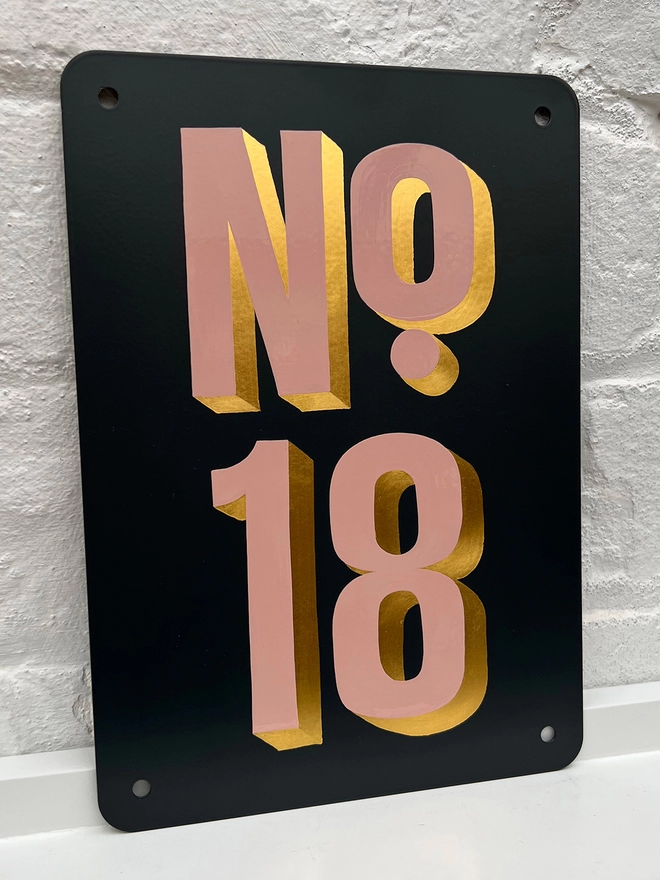 Dusky pink and gold leaf house number No.18, on anthracite grey metal plaque, against a white brick wall.