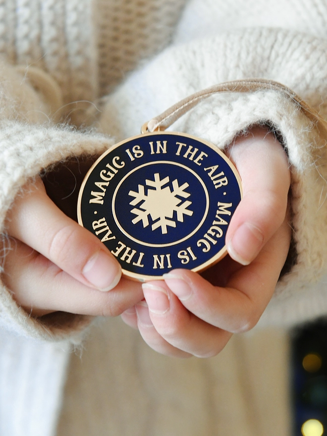 A navy blue and gold enamel Christmas decoration, with the words “Magic Is In The Air” surrounding a gold snowflake, is being held in two hands.