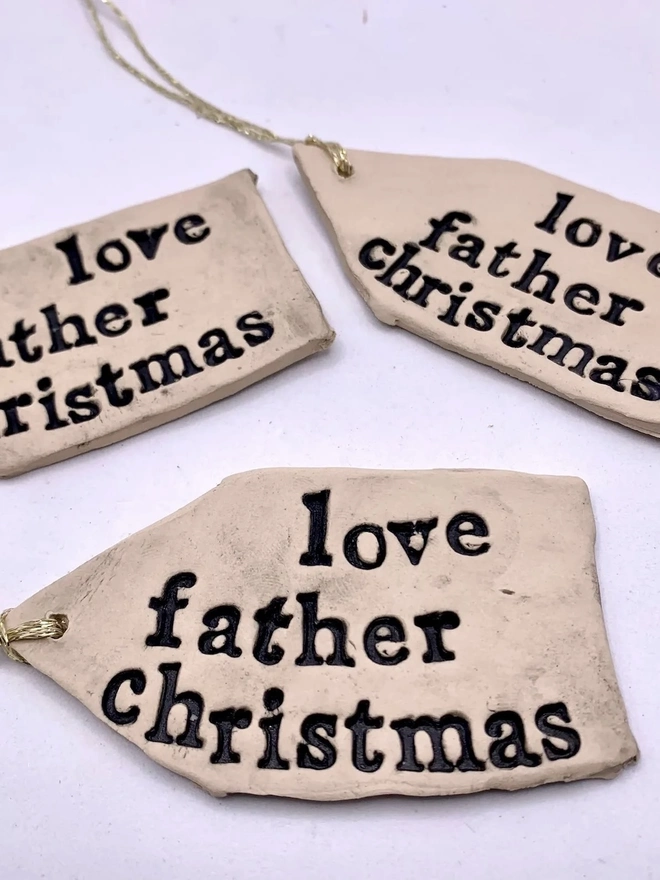 'Love Father Christmas' Handmade Ceramic Gift Tags, a group of ceramic gift tags attached sitting on a ale purple surface. 
