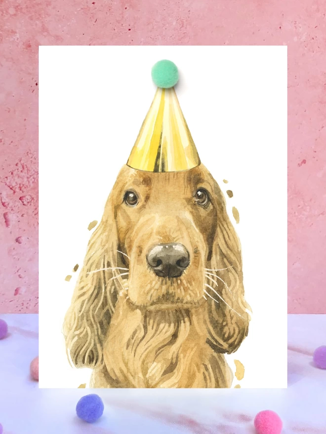 A greeting card featuring a hand painted design of a ginger cocker spaniel, stood upright on a marble surface surrounded by pompoms. 