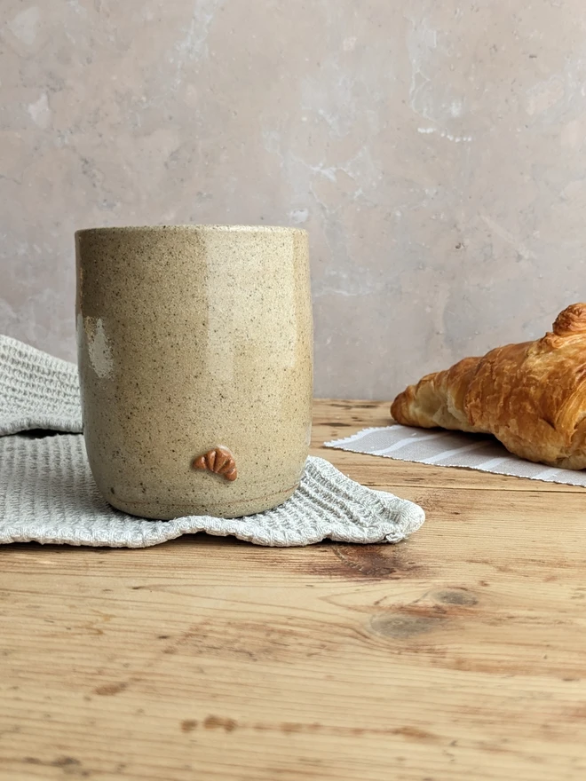 A biscuit-coloured croissant coffee cup with a subtle croissant design, placed against a grey background. 