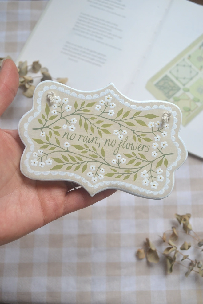 hanging clay sign with floral design reading no rain, no flowers' in front of gingham background