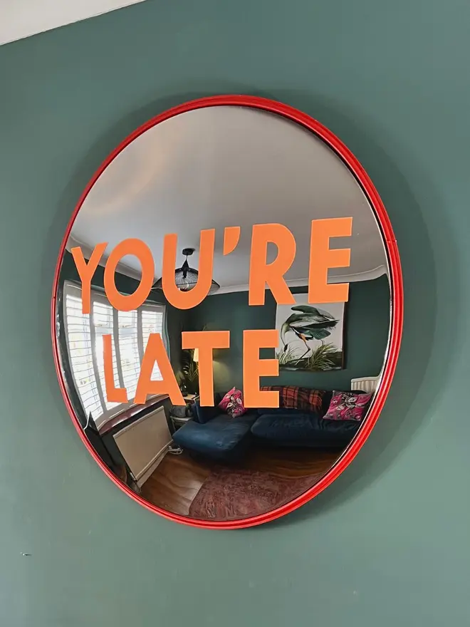 Round convex mirror with 'You're Late' hand painted on in orange, on a teal wall above a fireplace with a lamp in the corner