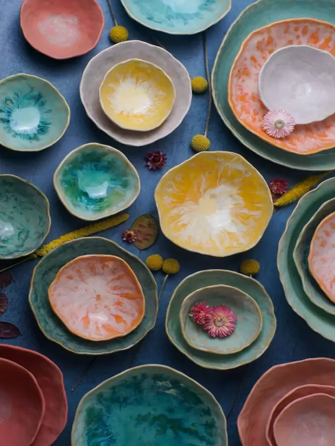 Selection of multicoloured ceramic nesting bowl, pottery bowls, serving bowls, citrus orange, yellow, green, turquoise, white, pink colours, breakfast bowl, dessert bowl, gift, homeware, kitchenware, tableware, photographed with flowers on a blue background