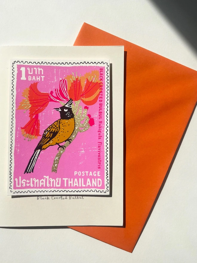 Bird Stamp Card Thailand Black Crested Bulbul, a white card with a colourful bird design in the centre on top of an orange envelope on a white surface. 