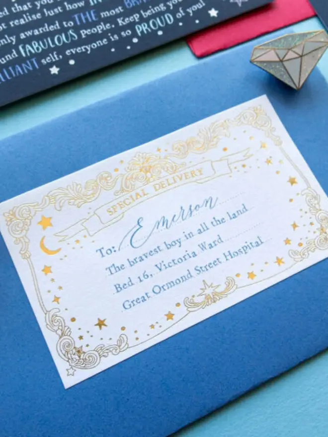 A close-up of a gold foil "Special Delivery" label on a blue envelope, addressed to Emerson, "the bravest boy in all the land." An enamel diamond pin and the navy certificate sit just above.