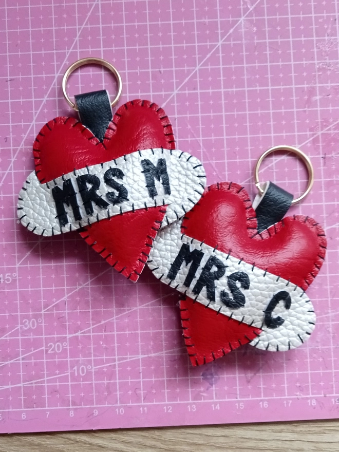 Two red leatherette tattoo style keyrings, with black lettering across white scrolls. One says MRS M, the other, MRS C