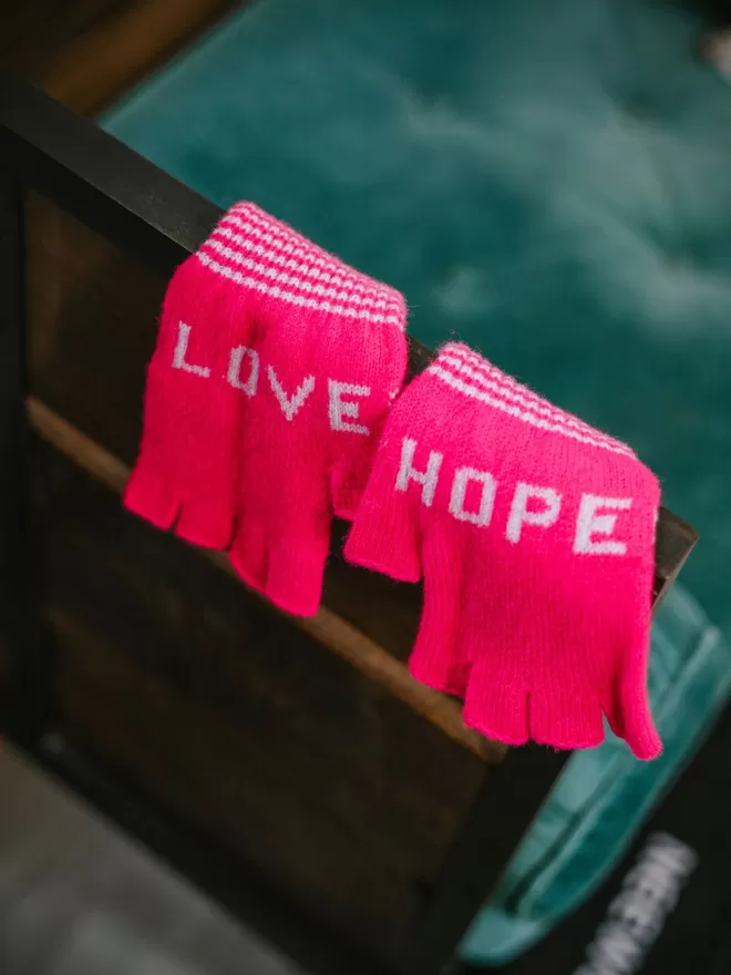 A bright pink fingerless knitted glove with the words LOVE and HOPE in lilac and a stripe cuff with orange edge detail. Made from 100% Lambswool with Wool spun in Yorkshire. 