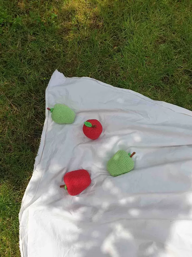Two green and two red crochet apples on a plain fabric with grass in the background.