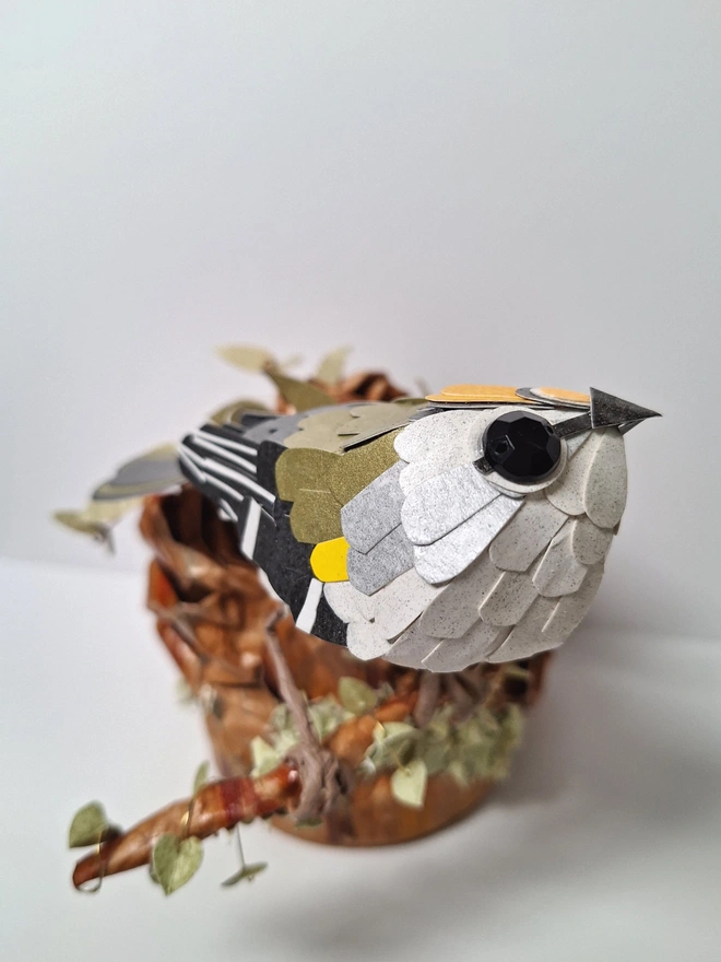 Firecrest paper bird sculpture, perched on a tree stump. lose up of the firecrest and its paper feathers.