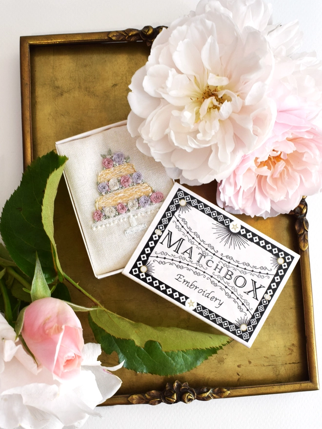 The finished embroidery placed inside the matchbox packaging for display.  Shown here on a vintage brass tray with blush pink and ivory roses cut from the garden.