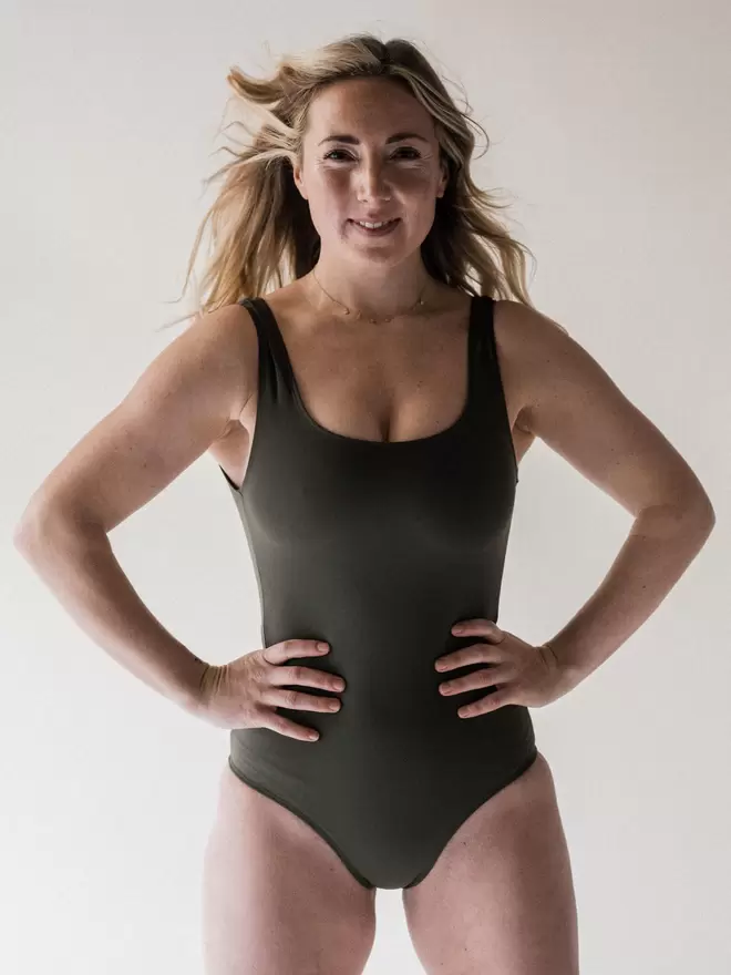 Smiling blonde woman in studio looking powerful with hands on hips wearing Davy J Sustainable Waterwear olive classic crossback swimsuit
