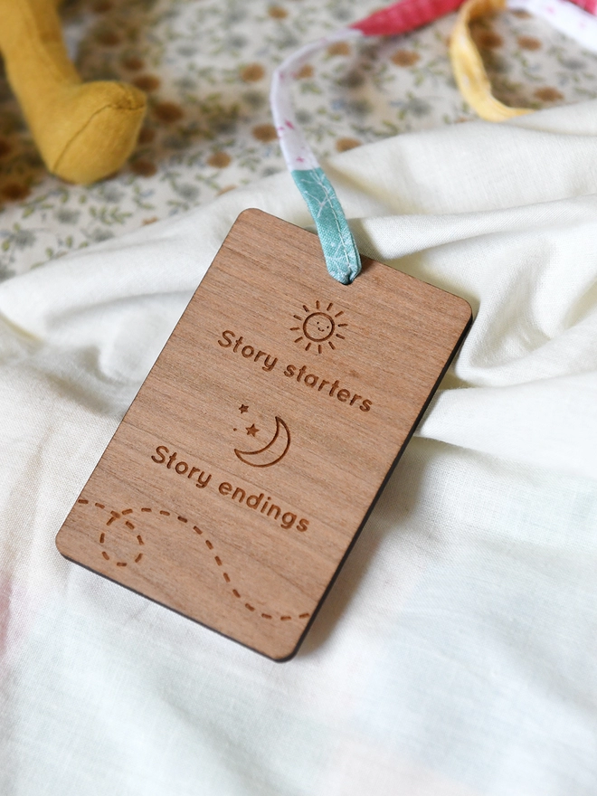 A wooden card is attached to a fabric ribbon. It has an engraved design showing a sun and a moon with the words Story starters and story endings.