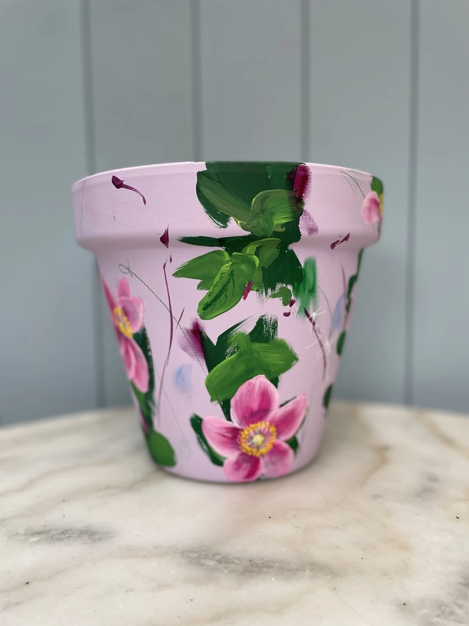 A beautifully hand painted interior terracotta plant pot. Background painted in a pale dusky pink adorned in pink Japanese anemone. 