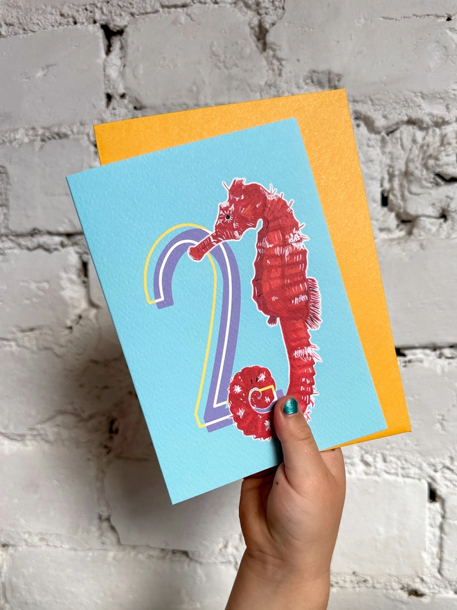 a bright blue card with a big purple number 2 and an illustration of a red seahorse and a bright yellow envelope