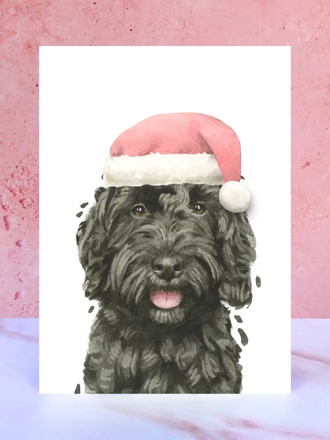 A Christmas card featuring a hand painted design of a black cockapoo, stood upright on a marble surface. 