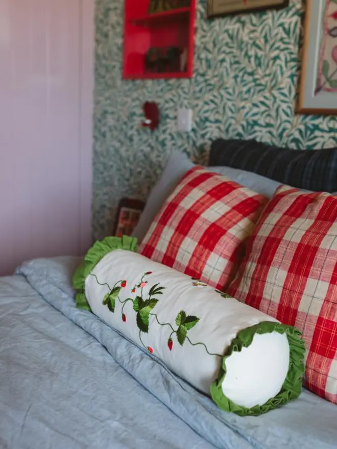 Embroidered strawberry bolster cushion in cream linen with green ruffle trim, styled on a colourful bed.