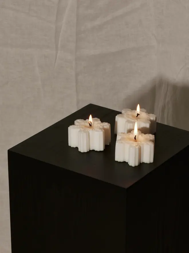 Classic Cream Snowflake Sculpture Candle. Three lit snowflake candles glowing on a black wooden plinth against a draped linen backdrop. The minimalist festive candles create a calm, elegant mood for winter evenings.