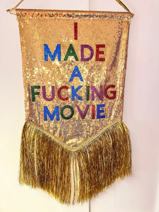 A mini customisable banner hangs from a hook. It has a gold sequin background and a gold tinsel trim along the bottom. The text is multicoloured and says 'I MADE A FUCKING MOVIE'