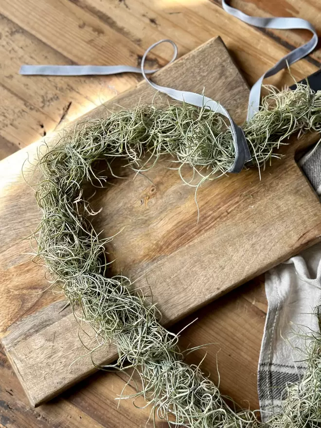 A wire Love Heart arrangement made of Tillandsia, finished  at the top with a thin ribbon for hanging, it lays across a wooden board, a ruffled vintage tea towel sits in the background along with a pair of small scissors.