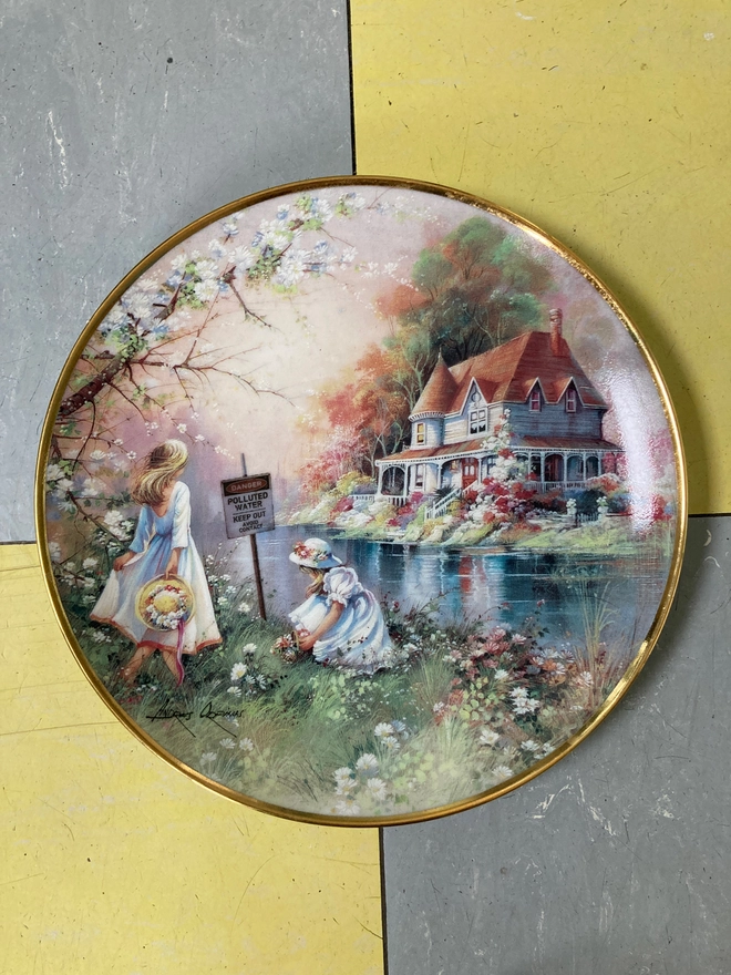 Beautiful decorative 8” plate featuring a watercolour style image of two young girls playing by a river. On the other side of the river is a beautiful white house. The scene is idyllic, yet in the river we see a warning sign which says ‘Danger! Polluted Water’...