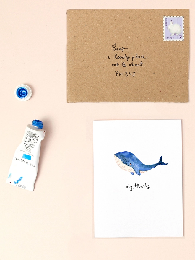 An image of a mini, rectangular white greetings card with an illustration of a blue whale with a hand written 'big thanks' message in black ink seen under the central illustration. The card is seen on top of a brown rectangular kraft envelope.