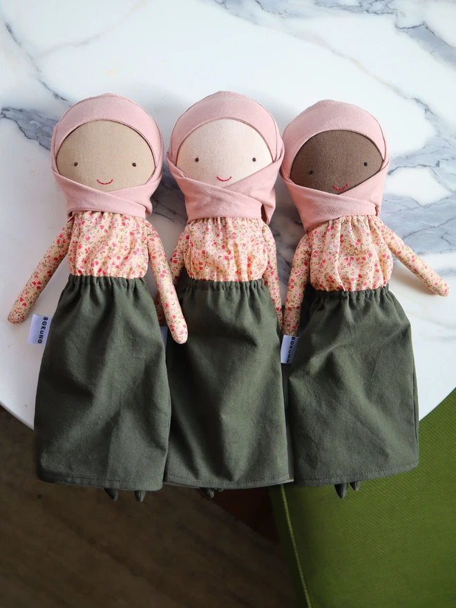 Three handmade cloth dolls wearing a blush pink hijab, a beige floral long-sleeve blouse, and an olive green skirt. The dolls have light, mid and dark fabric skin, simple stitched eyes and a small red smile, with a BOKUNO label on the side. 