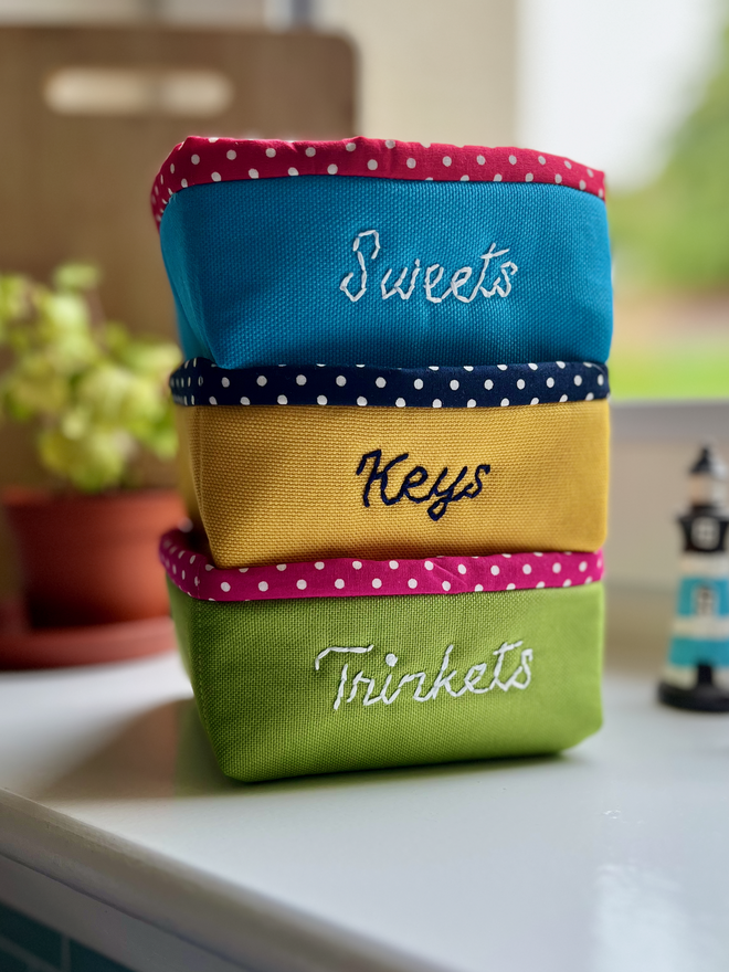 A stack of 3 fabric baskets on a windowsill, 1 green, 1 yellow, 1 blue, all with polka dot lining.