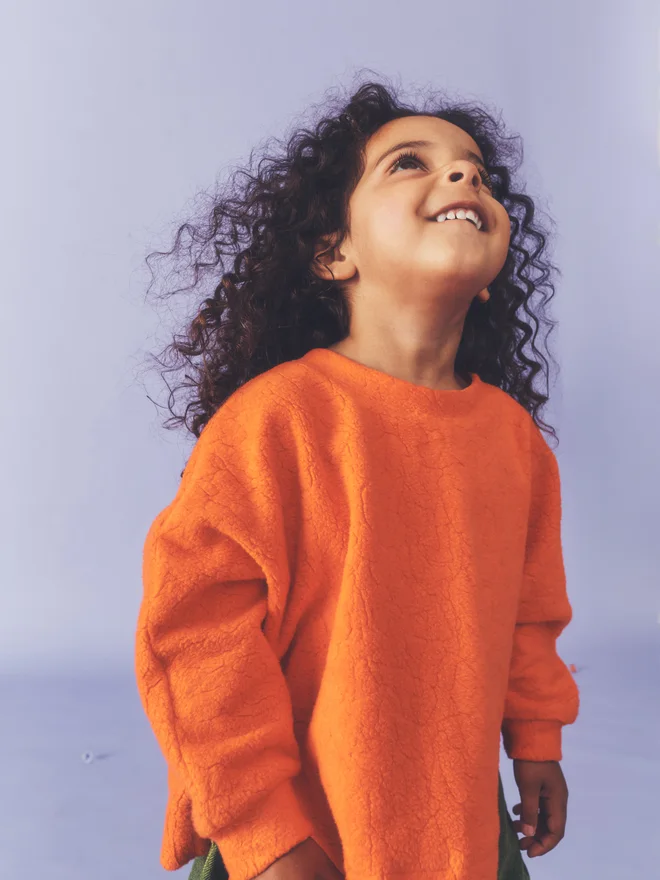 Close crop of a child smiling and looking up against a lilac backdrop, wearing a Studio Koter orange textured fleece top.