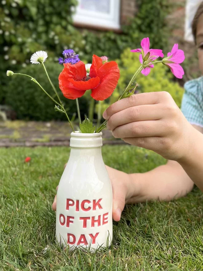 A child is arranging flowers in a bottle/vase, with ‘pick of the day’ lettering painted in red.