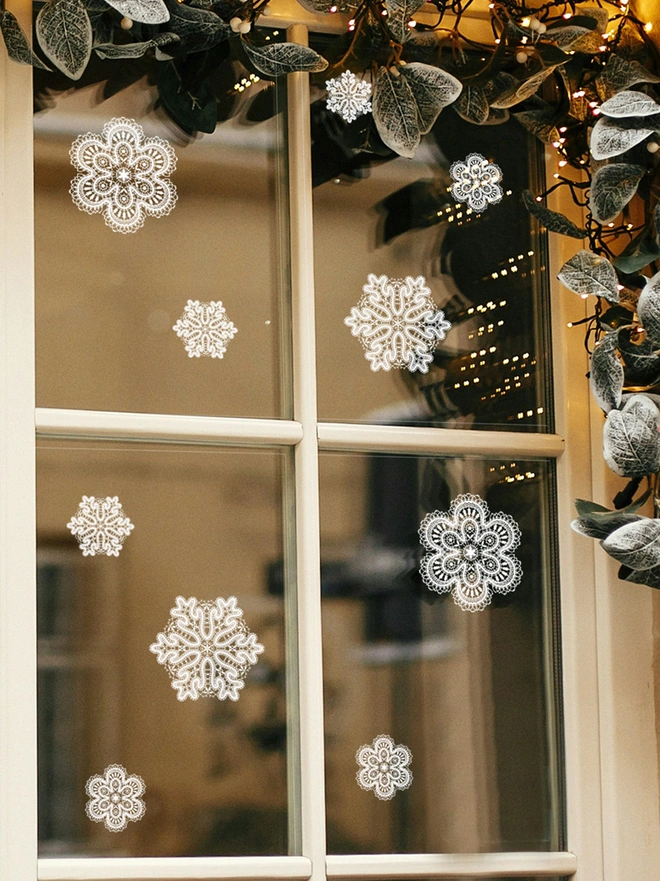 A window with a white frame and holly on the right hand side of the window with twinkly lights, on the window are 10 lace effect snowflakes of different sizes