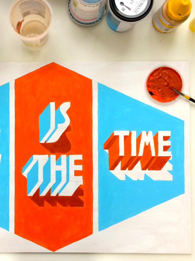 Painting of words Is The Time painted in 3d typography in orange and blue within a diamond shape by artist Survival Techniques. Paintbrush leans in a palette of orange paint with water pot and paint tubes above