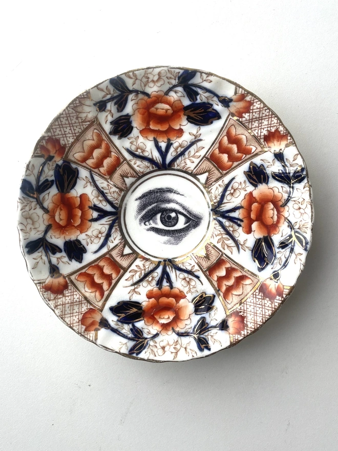 a really ornate plate with gold and orange florals and patterns, in the centre is printed a black and white illustration of an eye