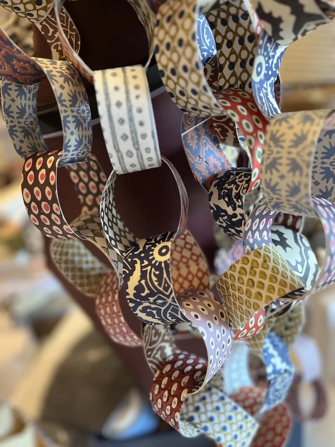 Colourful Paperchains (Set Of 60). Close-up of hanging paper chain garlands made from patterned links in navy, ochre, rust and red. The block-print-style designs make a colourful handmade decoration for festive or everyday styling.