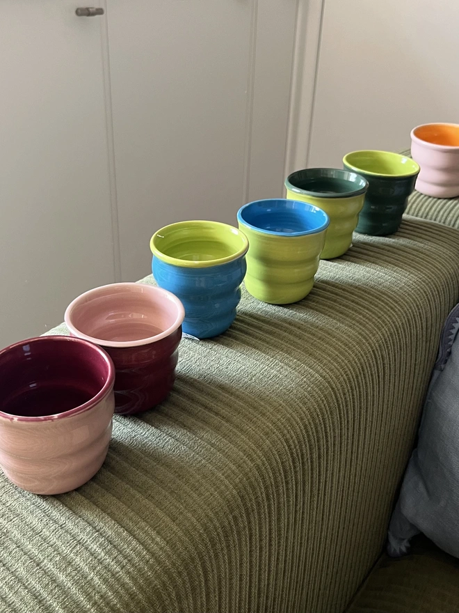 a row of colourful ceramic tumblers lined up on the arm of the sofa. the sofa arm is very square and wide and is made from an olive green cordurory fabric. 