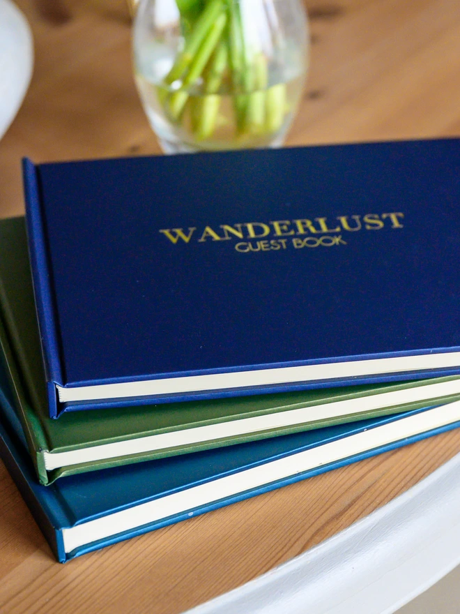 three guest books are piled on top of each other on a wooden table, The top one has been printed with the name of a house.