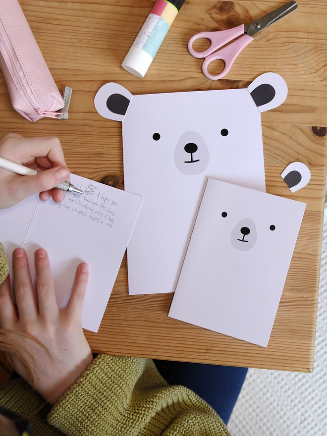 Several white polar bear greetings cards lay on a wooden table. A child is writing a message in one of the cards.