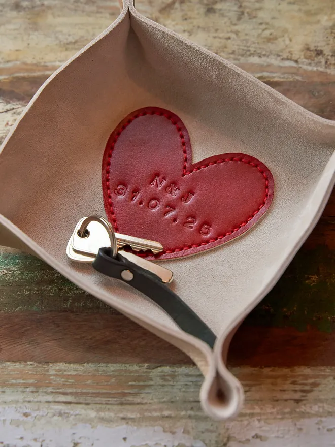 Personalised leather heart coin tray. A natural cream square leather tray with a red leather heart stitched to the base. The heart is personalised with a couples initials and their wedding date. Inside the tray sits a set of keys.