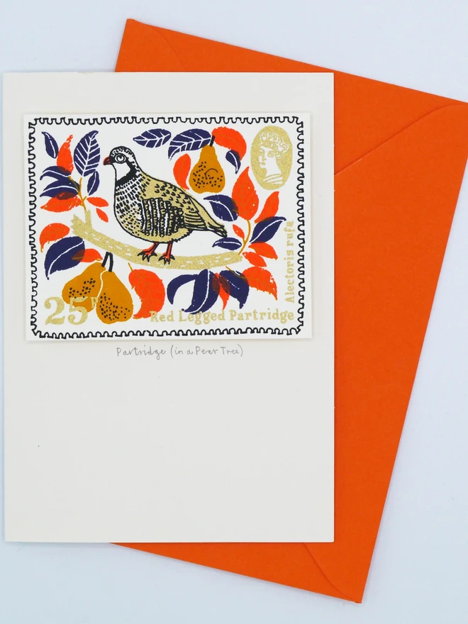 Partridge In A Pear Tree Christmas Card, a white card with a colourful bird design in the centre. It sits on top of an orange envelope on a plain surface. 