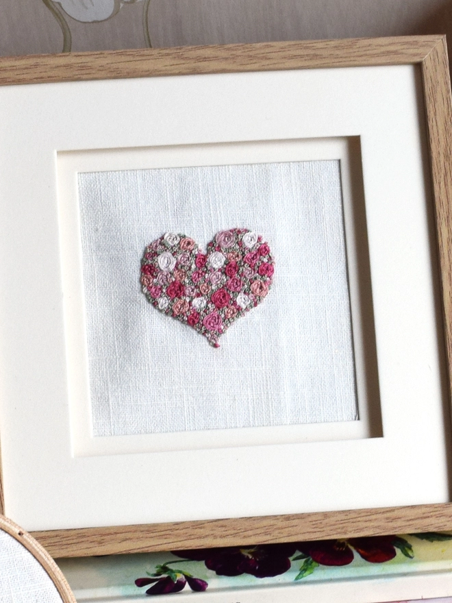 An embroidered Heart, of woven wheel roses in 5 shades of pink with French Knot green grass.  Displayed in a wooden Oak effect Frame with the glass removed. 