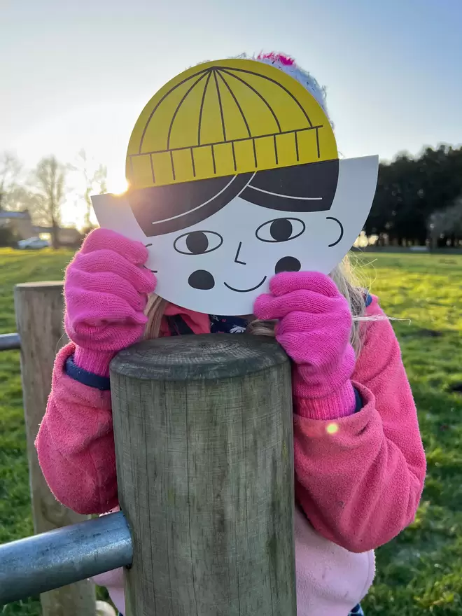 Child holding a round 'childs face' card in front of their face with yellow hat on.