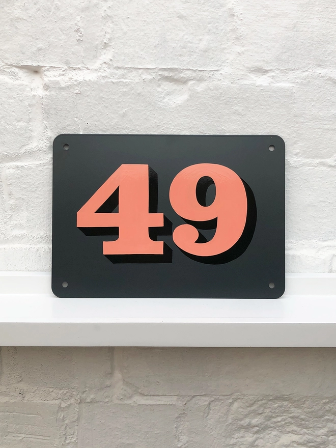 Hand painted house number against a white brick wall, number 49 in serif style in coral and black, on anthracite grey background. 