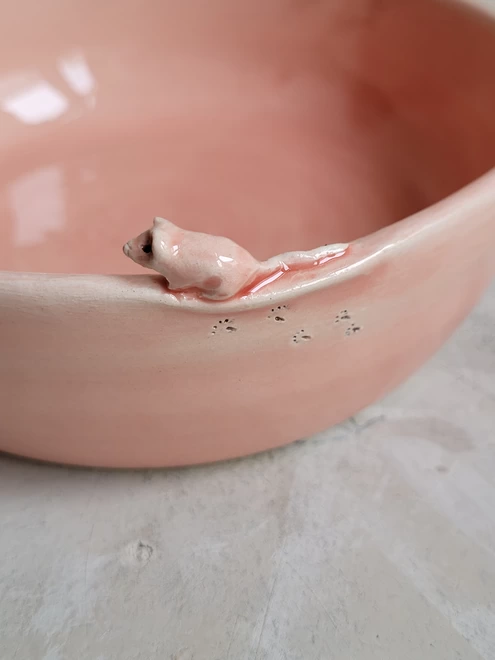 close up of a ceramic mouse attached to the rim of a pink pottery cat bowl there are foot prints etched into the clay on the side of the dish