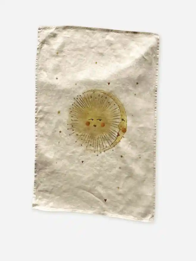 'Astral Weeks' Linen Tea Towel, a print sitting on a wooden surface with a sun motif in the centre. 